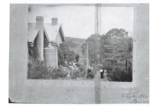 Black and white view of the side of Fernbank showing members of the Porter family, the water tank and the fruit produce of Strawberry Glen area in the background