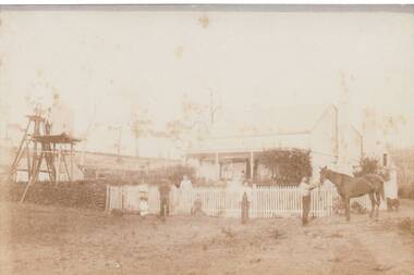 Weatherboard house, hipped roof, two chimneys, neat picket fence, drystone wall on left in fromt of windmill, family and horse in front.