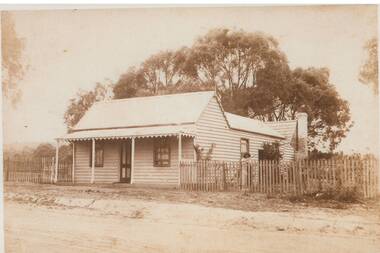 Single story wooden cottage, at front central door, window either side, plain verandah and picket fence, house extends back to chimney, large tree behind.