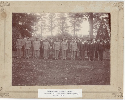 Two rows of 10 men standing to attention with rifles, left front seven in uniform, rest in suits. Trees and bystanders behind.