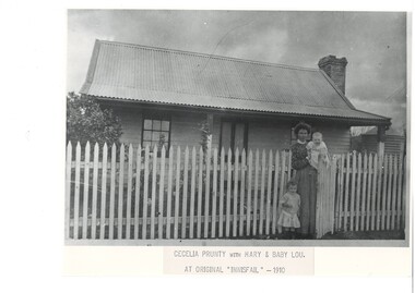 Black and white photograph of Cecelia Prunty with daughters Mary and Baby Lou at the front gate at "Innisfail" in 1910. The corrugated iron roof dominates the image while parts of the garden and verandah are visible through the picket fence