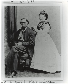 Studio photograph, Victorian couple, bearded man in suit seated left, woman in white dress standing right.