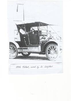 Early two-seater car, soft hood, lady behind the wheel, in front of wooden building.