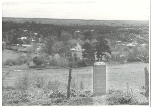 View from mount looking down on township, heavily treed, glimpses of houses, church clear in centre, simple stone monument in foreground.