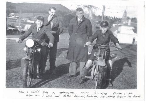 Roadside, Two boys in school uniforms seated on motor-bikes, two men between them in winter coats. Shops, some houses and Mount in background.