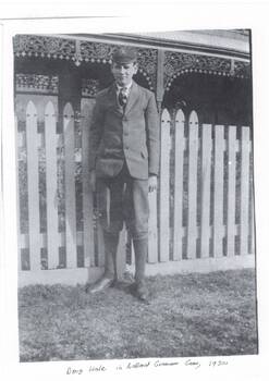 Boy in school uniform standing in front of house with iron lace veranda and picket fence. Uniform is blazer, cap, tie knickerbockers, long socks.