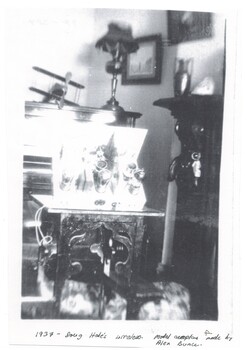 Living room, showing part of upright piano with lamp and model plane, homemade wireless front centre, Edge of fireplace on right with vase, pictures on walls.