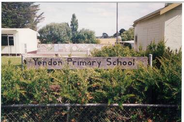 This colour photograph shows part of two of the school buildings and a mural while the foreground features native plants and the sign "Yendon Primary School" on the school fence.