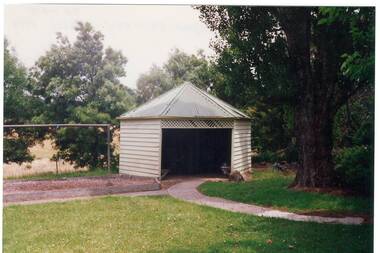 A coloured photograph of a six sided shelter shed in the grounds of the former Yendon Primary School surrounded by trees, lawn and pathways