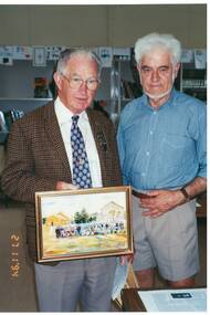 Two men, one in jacket and tie, the other in shirtsleeves and shorts, holding framed picture of people and church.