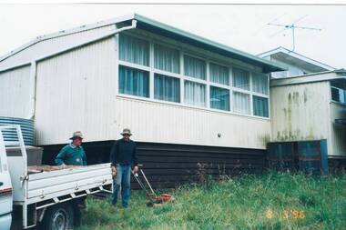 View of portable buildings of Clarendon school prior to their sale, two men with mower and vehicle in foreground.