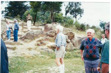 Group of people on sloping ground, boulders and grass, steps on left, trees behind