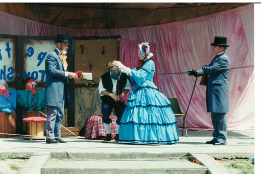 On stage, lady in blue crinoline crowning bearded man, flanked by other men all in Victorian dress.