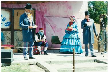 Four actors in Victorian dress, beaded gent seated centre in shirtsleeves and vest, to his left lady in blue crinoline and bonnet, holding crown on a cushion, either side gents in top hats and frock coats.