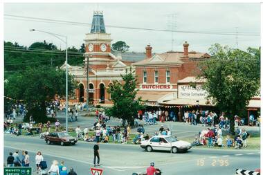 Street view, brick town hall, butchers shop, electrical store, many people milling around.