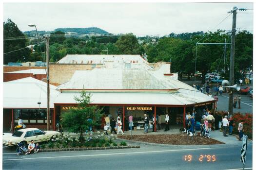 View down onto street corner, antique shop with verandah, mount on horizon, many parked cars and people..