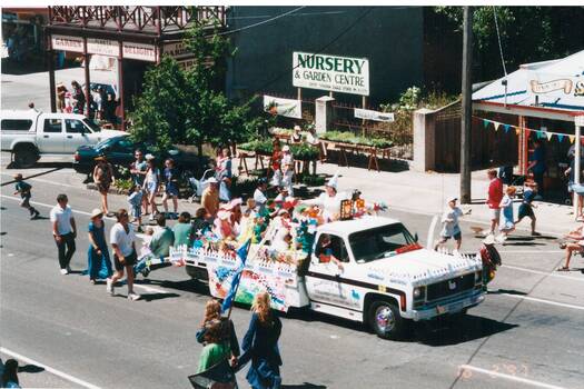 Street procession, decorated white ute, children seated in tray, adults walking beside and behind.