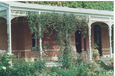 Section of brick house with iron lace veranda, partly covered with ivy, climbing roses and jasmine.