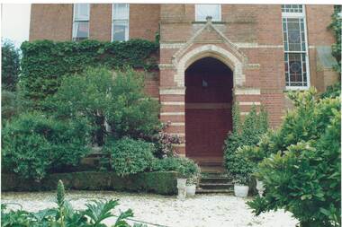 Double story section of polychrome brick house, part covered in ivy, lush garden, stained glass window, steps up to arched porch.