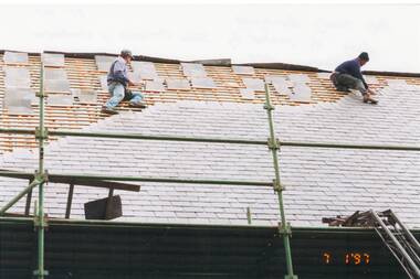 Two workmen replacing slate shingles on church roof, partly completed, roof struts exposed.