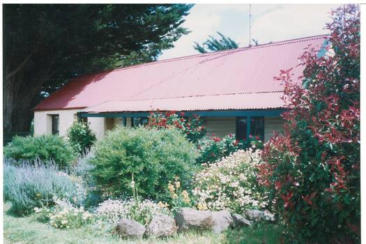 Long, low cream rendered building, low pitched red iron roof and verandah, lush garden.