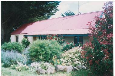Long, low cream rendered building, low pitched red iron roof and verandah, lush garden.