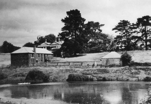 View uphill across ornamental lake, bluestone brewery with house behind, tall trees on top of rise.