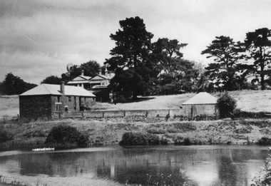 View uphill across ornamental lake, bluestone brewery with house behind, tall trees on top of rise.