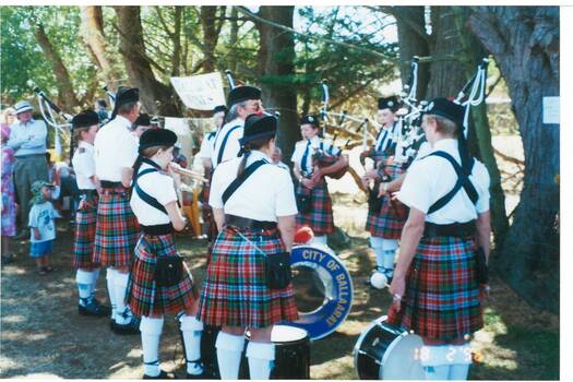 Six young men in kilts, from behind, under trees, two are holdong drums.