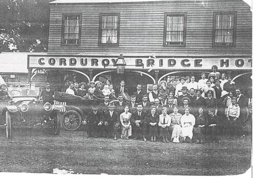 Two story wooden building, 4 windows on upper floor, sign across veranda "Corduroy Bridge Hotel, weel dressed crown seated on benches and in two early cars.