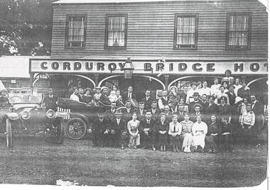 Two story wooden building, 4 windows on upper floor, sign across veranda "Corduroy Bridge Hotel, weel dressed crown seated on benches and in two early cars.