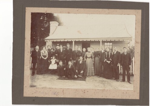 Family portrait, 21 well-dressed men, women and children, in front of single story 2 room cottage with veranda with fretwork fascia