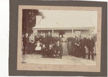 Family portrait, 21 well-dressed men, women and children, in front of single story 2 room cottage with veranda with fretwork fascia