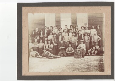 Some 50 men and youth, most standing, some crouching, some reclining in rough rows before a wooden building with tall windows, all in victorian workers clothes.