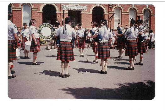 Pipe band, wearing kilts, on paved forecourt of brick building, arched windows and openings.