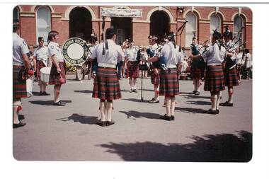 Pipe band, wearing kilts, on paved forecourt of brick building, arched windows and openings.