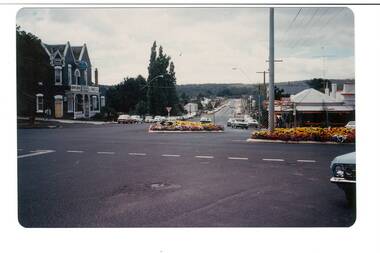View across intersection, little traffic, hotel on left, shops on right, trees centre.