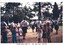 Children in foreground watching pipe band in park, tall cypress trees