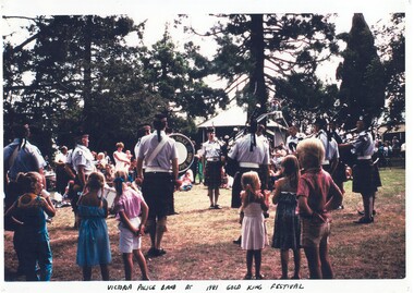 Children in foreground watching pipe band in park, tall cypress trees