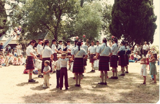 Children  watching pipe band in clearing in park, tall trees behind.