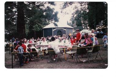 Crowd seated at lunch in park, rotunda behind, surrounded by trees.