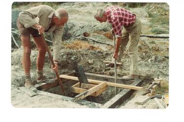 two  men in shirtsleeves digging out foundation hole in wooden frame