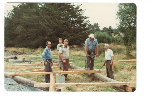 Men working on construction, wooden frames and logs on ground, trees in background
