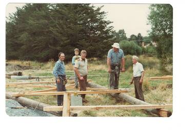 Men working on construction, wooden frames and logs on ground, trees in background
