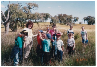 Group of children and adult in grassy field, few trees in background.