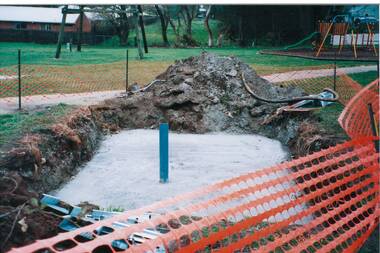 In a park, close up of rectangular concrete slab in ground, blue pipe extending upwards, 