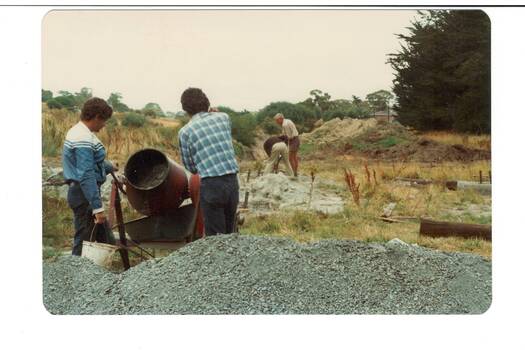 Construction area in park, very rough ground, two men operating concrete mixer, men digging in background