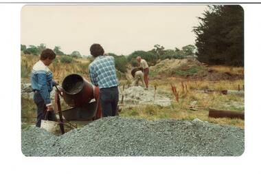 Construction area in park, very rough ground, two men operating concrete mixer, men digging in background