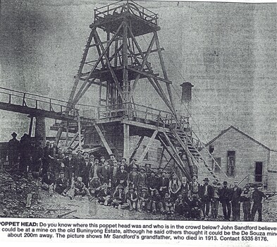 Black and white image of a mine poppet head and a large group of miners at the base of the structure. Several other buildings and a chimney can be seen in the background.