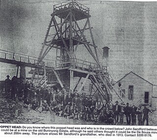 Black and white image of a mine poppet head and a large group of miners at the base of the structure. Several other buildings and a chimney can be seen in the background.